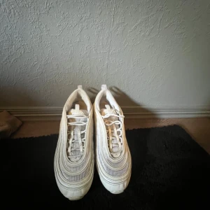 Nike Air Max 97 vita sneakers - Nike Air Max 97 sneakers i vitt med ikoniska vågiga linjer och synlig Air-sula. Skorna har snörning och en rund tå, med detaljer i syntet och mesh för en sportig vibe. Perfekt för dig som gillar klassisk streetstyle och vill ha en clean look.