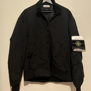 Svart bomberjacka från Stone Island - Svart bomberjacka från Stone Island med klassisk ribbad krage och muddar. Jackan har dragkedja framtill, två sidofickor och den ikoniska Stone Island-loggan på ärmen. Materialet är polyester och jackan har en loose passform som ger en chill vibe.