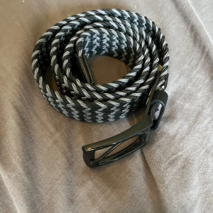 Svart och grått flätat bälte - Snyggt flätat bälte i svart och grått med stilren metallspänne. Bältet har ett coolt mönster och är gjort av syntetmaterial med detaljer i svart skinn vid spännet. Perfekt accessoar för att lyfta din outfit. Pris ej hugget i sten vid snabb affär. hör av dig vid minsta fundering!
