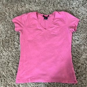 Rosa v-ringad t-shirt från Gant - Snygg rosa t-shirt från Gant med v-ringning och broderad logga på bröstet. Klassisk passform och korta ärmar, perfekt till jeans eller shorts. Tillverkad i mjuk bomull som känns skön mot huden.