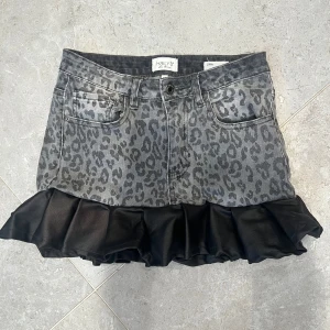 Leopardmönstrad jeanskjol med volang - Cool kort jeanskjol från Haily's med grått leopardmönster och bred svart volangkant nedtill. Kjolen har klassiska fickor fram och bak samt bälteshällor. Perfekt statement-plagg för dig som gillar att sticka ut.