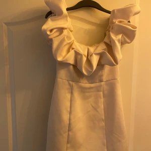 Champagnefärgad kort klänning med volang - Superfin kort klänning i champagnefärg med stora volangdetaljer runt axlarna. Klänningen har en elegant och modern look med en figurnära passform och är tillverkad i ett glansigt material som ger en lyxig känsla. Perfekt för dig som vill sticka ut. Funkar perfekt till studentfirande, troförlovning, förlovning, bröllop och andra festliga tillfällen.