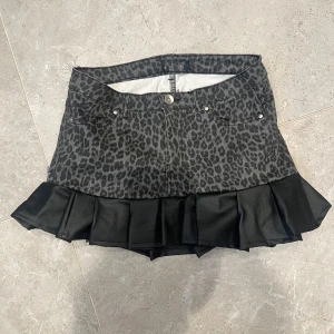 Leopardmönstrad kortkjol med volang - Snygg kortkjol med svart leopardmönster och bred volangkant i satinliknande material. Kjolen har klassisk jeansmodell med fickor fram och bak samt knapp och dragkedja. Perfekt statement-plagg för dig som vill sticka ut.