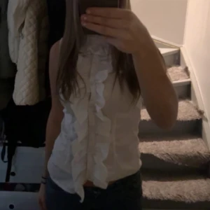 Vit ärmlös blus med volang - Supersöt vit blus med volangdetaljer längs hela framsidan. Ärmlös modell med hög hals och lätt figursydd passform. Perfekt för dig som gillar en romantisk och trendig look.