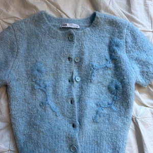 Blå kofta med broderade blommor Zara - Superfin ljusblå kofta från Zara med kort ärm och broderade blommor framtill. Koftan är stickad i mjukt material och har knappar hela vägen. Perfekt för lager-på-lager och ger en söt touch till din outfit.