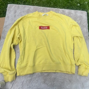 Gul sweatshirt från NA-KD - Gul sweatshirt från NA-KD med röd patch och vit brodyr på bröstet. Tröjan har rund hals, ribbade muddar och är långärmad. Perfekt för dig som gillar färg och vill sticka ut lite extra. Materialet är mjuk bomull som känns skönt mot huden.