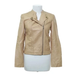 Beige skinnjacka från 365, stl 36 - Snygg beige skinnjacka från 365 med coola detaljer som sned dragkedja, markerade axlar och quiltade partier vid axlar och nederkant. Jackan har två fickor framtill och en stilren, figurnära passform. 