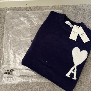 Mörkblå stickad tröja från Ami - Säljer en mörkblå stickad tröja från Ami Alexandre Mattiussi med deras ikoniska vita hjärta och A-logga på bröstet. Tröjan har rund halsringning och är i mjukt material som känns lyxigt och skönt. Perfekt för dig som gillar stilrena detaljer och vill ha något trendigt.