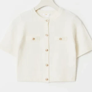 Vit kortärmad kofta med knappar - Superfin vit kofta med korta ärmar och snygga guldfärgade knappar framtill. Tröjan har två dekorativa fickor på bröstet och är stickad i ett mjukt material. Perfekt för lager-på-lager och ger en clean look.