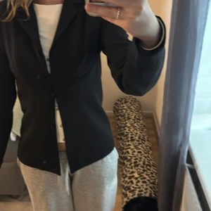 Svart kavaj från NA-KD, storlek 36 - Snygg svart kavaj från NA-KD i figurnära modell med klassisk krage och knappar framtill. Jackan har långa ärmar och är tillverkad i ett slätt, lätt glansigt material som ger en stilren look. Perfekt att styla med jeans eller kjol för en trendig outfit. Bygel som ska uringa bröstkorgen. Har aldrig använt. Pris går att diskutera.