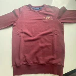 Sweatshirt från GANT - Vinröd sweatshirt från GANT med broderad logga på bröstet. Klassisk rund halsringning och lång ärm. Mjuk bomullskänsla och stilren design som passar perfekt till jeans eller chinos.
