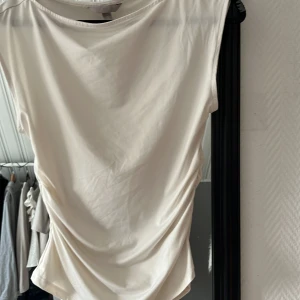 Vit omlottopp med rynk från H&M - Säljer en vit omlottopp från H&M i mjukt material. Toppen har ärmlös design och snygga rynkade detaljer på sidan som ger en cool draperad effekt. Perfekt att styla med jeans eller kjol för en fräsch look.
