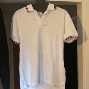 Vit pikétröja från Prada med röd detalj - Snygg vit pikétröja från Prada med klassisk krage och knäppning framtill. Tröjan har röda och svarta ränder på krage och ärmslut samt diskret Prada-logga på bröstet. Tillverkad i bomull för en skön och fräsch känsla. Perfekt för en clean och stilren look.