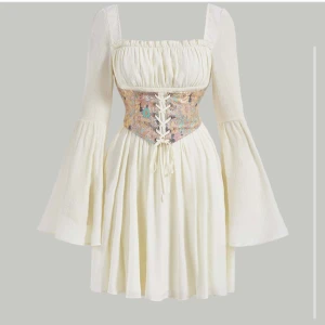 Vit kortklänning med blommig snörkorsett - Superfin vit kortklänning med vida långa ärmar och fyrkantig ringning. Midjan markeras av en broderad blommig korsett med snörning framtill i pastelliga färger. Klänningen har flowy kjol och romantiska vibbar, perfekt för dig som gillar boho-stil. 🌸 passar storlek M-L