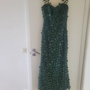 Grön långklänning med 3D-blommor - Magisk grön långklänning med transparent mesh upptill och massor av 3D-blommor och glitterdetaljer över hela klänningen. Figurnära passform upptill och utsvängd nertill, perfekt för bal eller fest. Klänningen har ärmlös design och vacker illusion-neckline.