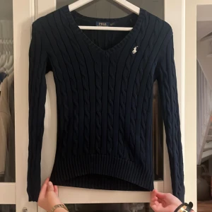 Mörkblå kabelstickad tröja Polo Ralph Lauren - Kabelstickad mörkblå tröja från Polo Ralph Lauren med v-ringning och broderad logga på bröstet. Tröjan har lång ärm, ribbade muddar och en slim passform som sitter snyggt. Perfekt för dig som gillar klassisk och stilren look.