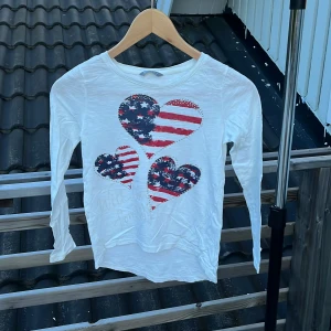 Vit långärmad tröja med hjärttryck Lindex - Vit långärmad tröja från Lindex med tre stora hjärtan framtill i rött, blått och vitt med stjärnor och ränder. Tillverkad i mjuk bomull, perfekt för dig som gillar coola tryck och bekväma plagg. Rund halsringning och normal passform.