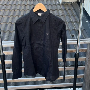 Svart randig skjorta från Esprit - Snygg svart skjorta från Esprit med diskreta ränder och klassisk krage. Skjortan har lång ärm, knäppning framtill och liten broderad logga på bröstet. Perfekt för dig som gillar stilrena och enkla plagg med en twist.