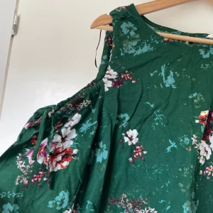 Grön blommig blus med knytärm - Supersöt grön blus med blommigt mönster i vitt, rosa och rött. Har en offshoulder look fast långarmad.