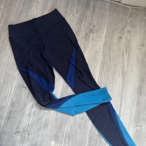 Nike Dri-Fit leggings blå och svart S - Nike Dri-Fit leggings i svart med blå och turkosa detaljer. Hög midja och tight passform, perfekta för träning. Tillverkade i ett syntetmaterial som andas och transporterar bort fukt. Snygg design med färgblock längs benen. Ytludd finns enligt bilder.