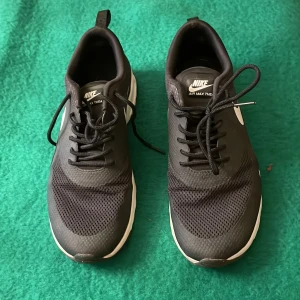 Nike Air Max Thea svarta sneakers - Svarta Nike Air Max Thea sneakers med vit sula och klassisk vit swoosh på sidan. Ovandelen är i mesh och syntet för en sportig look och skön känsla. Skorna har snörning och en låg profil som passar till streetwear och träning.