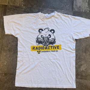 Radioactive Chernobyl vit t-shirt - Vit t-shirt med tryck av en panda med gasmask och texten 'RADIOACTIVE' i gult och svart. Under står det 'chernobyl-tour.ua'. Klassisk passform med rund hals och korta ärmar. Perfekt för dig som gillar unika och grafiska motiv.