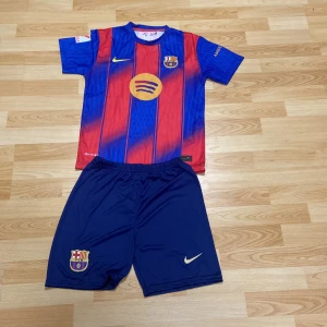 FC Barcelona Lamine Yamal matchställ - FC Barcelona matchställ med Lamine Yamal 19-tryck. Tröjan är kortärmad med klassiska blå och röda ränder, klubbmärke och Nike-logga. Materialet är lätt och andas, perfekt för fotboll. Shortsen är marinblå med klubbmärke och Nike-logga.