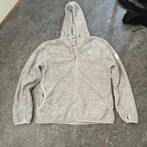Grå teddyjacka med huva från The Peak - Mysig grå teddyjacka från The Peak i storlek L. Jackan har huva, dragkedja framtill, två fickor och tumhål vid ärmsluten. Perfekt för kalla dagar när du vill vara både varm och chill. Fleece-liknande material som känns riktigt skönt mot huden.