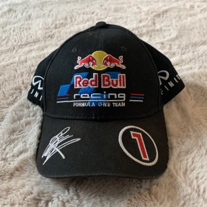 Red Bull Racing keps - Svart keps från Red Bull Racing med broderade loggor, nummer 1 och Infiniti-detaljer. Kepsen har färgglada broderier i rött, blått, gult och vitt samt en autograf på skärmen. Perfekt för motorsportfans som vill sticka ut.