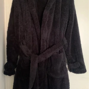 Svart teddy/ morgonrock med bälte - Mysig svart teddy/ bad / morgonrock med bälte i midjan och stora fickor. Morgonrocken har långa ärmar med knappar vid ärmsluten och en fluffig, oversized känsla andvänd ett fåtal gånger