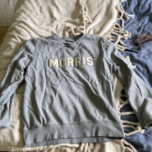 Grå sweatshirt från Morris Gents - Grå sweatshirt från Morris Gents med broderad logga i vitt framtill. Klassisk rund halsringning, ribbade muddar och lång ärm. Perfekt för en chill och stilren look. Passar för både tjej eller kille! 