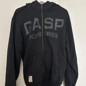 Svart hoodie från GASP Industries - Svart hoodie från GASP Industries med dragkedja och stor logga framtill. Huva med orange broderad detalj på baksidan. Mjuk bomullskänsla, känguruficka och ribbade muddar. Perfekt för dig som gillar sportig och avslappnad stil.