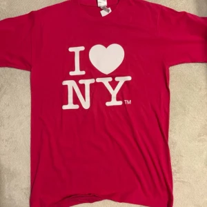 Rosa I Love NY t-shirt - Säljer en klassisk rosa t-shirt med det ikoniska vita 'I ♥ NY'-trycket på bröstet. T-shirten har rund hals och korta ärmar, perfekt för dig som vill visa lite New York-vibes. Materialet känns mjukt och skönt mot huden.