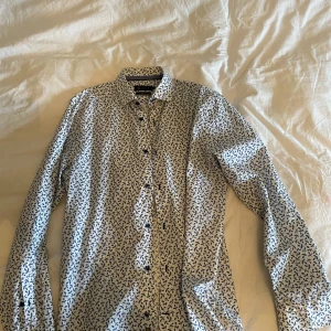 Vit skjorta med blått mönster - Snygg vit skjorta från Riley med blått abstrakt mönster över hela tyget. Köp denna skjortan för 100 kr och få med den andra skjortan gratis på köpet eller denna separat för 80 kr! 