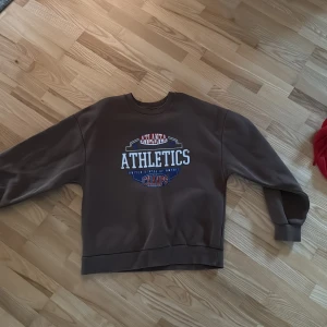 Brun sweatshirt med Athletics-tryck - Säljer en brun sweatshirt med stort Athletics-tryck i vitt, blått och rött på bröstet. Tröjan har rund hals, långa ärmar och en loose passform. Perfekt för en sportig och avslappnad stil.