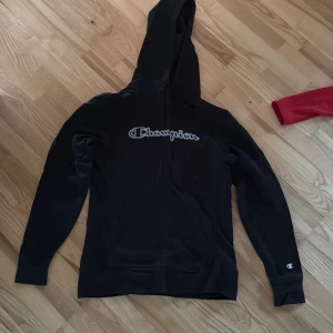 Svart Champion hoodie med huva - Svart hoodie från Champion med stor logga broderad på bröstet och klassisk känguruficka. Tröjan har huva med dragsko och långa ärmar med Champion-logga på mudden. Perfekt för en chill och sportig stil.