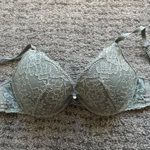 Grå/grön bh - Snygg grå bh från H&M i storlek 75C, täckt med blommig spets och vadderade kupor. Axelbanden är justerbara och framtill finns en liten dekorativ detalj. Endast använd några fåtal gånger. 
