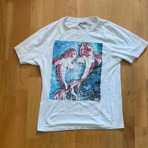 Vit t-shirt med fisktryck från Jacart - Vit t-shirt från Jacart med ett stort färgglatt tryck av två fiskar i blått och rött på framsidan. Modellen är relaxed fit och har korta ärmar. Perfekt för dig som gillar unika och konstnärliga motiv. Gjord i mjuk bomull för skön känsla.