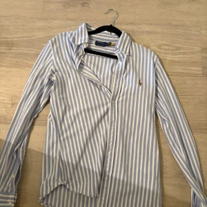 Randig skjorta från Polo Ralph Lauren - Klassisk skjorta från Polo Ralph Lauren i ljusblått och vitt med vertikala ränder. Skjortan har lång ärm, knappar framtill och broderad logga på bröstet. Tillverkad i mjuk bomull och har en tidlös, normal passform.