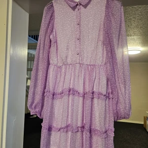 Ljuslila volangklänning med krage - Supersöt ljuslila klänning med små vita mönster, volanger och knappar framtill. Klänningen har lång ärm, klassisk krage och  lager med volangdetaljer. Perfekt för dig som gillar romantisk och lekfull stil.