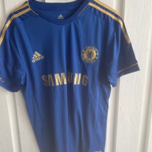 Chelsea Hazard 17 matchtröja Adidas - Chelsea FC fotbollströja med Hazard 17 på ryggen. Klassisk blå färg med guldiga detaljer och Adidas-logga. Tryck med Samsung som sponsor framtill och klubbmärke på bröstet. Korta ärmar och tillverkad i lätt, ventilerande material.