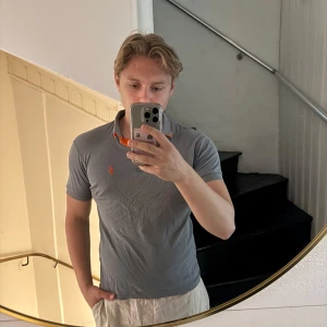 Grå pikétröja från Polo Ralph Lauren - Säljer en grå pikétröja från Polo Ralph Lauren i storlek M. Tröjan har klassisk krage, korta ärmar och orange logga på bröstet. Insidan av kragen är orange för en snygg kontrast. Perfekt för en clean och sportig look.