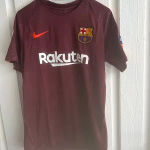 FC Barcelona Messi vinröd matchtröja - Snygg FC Barcelona fotbollströja från Nike i vinröd färg med orange detaljer. Tryck med Messi 10 på ryggen och Rakuten-logga på bröstet. Kortärmad modell med klubbmärke och Champions League-märke på ärmen. Perfekt för dig som älskar Barca och Messi.
