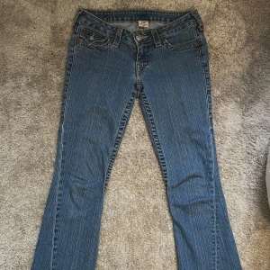 True Religion blå bootcut jeans - Säljer ett par klassiska blå jeans från True Religion med bootcut passform. Jeansen har snygga kontrastsömmar, ikoniska bakfickor med lock och silverfärgade knappar. Tillverkade i USA i slitstarkt denim med orange detaljer vid sömmarna. Dem hadde slitage vid övre delen av fickorna men dem är professionellt fixade och syns förövrigt inte så mycket🤗