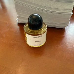Byredo 1996 Eau de Parfum - Byredo 1996 Eau de Parfum i en rund glasflaska med svart rund kork. Parfymen har en guldfärgad vätska och minimalistisk vit etikett med svart text. En stilren och modern doft från ett populärt svenskt parfymhus. Helt ny och oanvänd! 