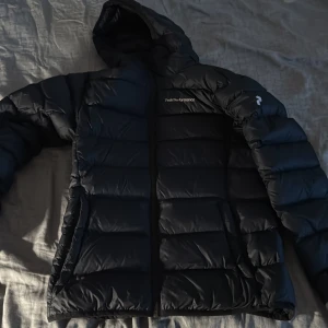 Svart dunjacka Peak Performance S - Säljer en svart dunjacka från Peak Performance i storlek S. Jackan har huva, dragkedja framtill och klassisk pufferdesign med breda kanaler. Perfekt för kalla dagar och riktigt snygg till streetwear-looken.