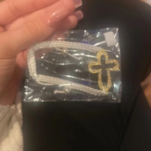 Vitt armband med guldfärgat kors - Snyggt armband med flätad vit tråd och justerbar knytning. Armbandet har ett stort guldfärgat kors med glittriga detaljer som ger extra bling. Perfekt accessoar för dig som vill sticka ut med något unikt och trendigt.