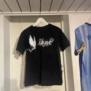 Svart Vlone t-shirt med tryck - Svart t-shirt från Vlone med stort vitt V-tryck på ryggen och coolt motiv med text och rökdetaljer framtill. Skön passform och klassisk rund hals. Perfekt för dig som gillar streetwear och vill sticka ut med en unik design.