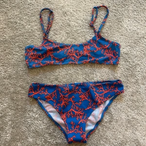 Blå bikini med korallmönster - Snygg bikini i blått med rött korallmönster. Toppen har smala axelband och raka kanter, och bikinitrosan har klassisk passform. Perfekt för stranden eller poolen, med färgstark och unik look. Skriv om du vill köpa överdel/underdel separat!