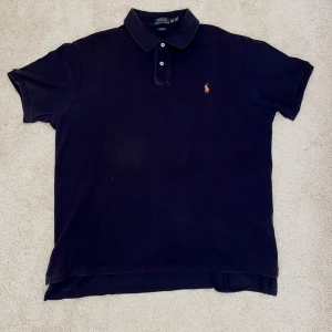 Blå pikétröja Polo Ralph Lauren  - Klassisk blå pikétröja från Polo Ralph Lauren i storlek XXL. Tröjan har röd broderad logga på bröstet, krage och knäppning med två knappar. Custom fit-modell i mjuk bomull som är både snygg och bekväm.